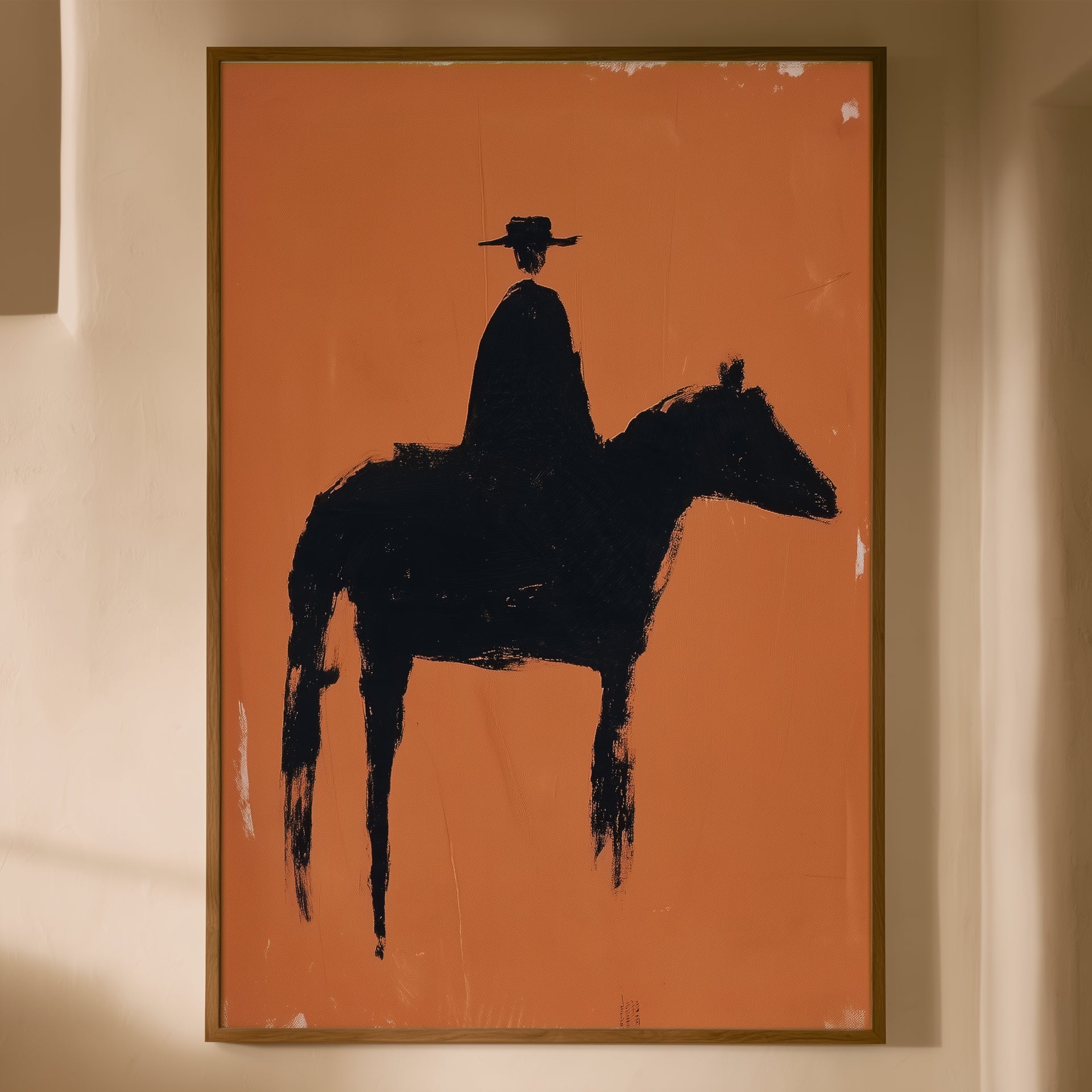 A framed art print featuring a bold black silhouette of a cowboy riding a horse against a solid orange background, hung on a warm beige wall.